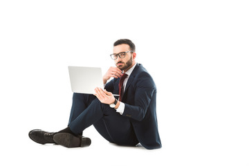 thoughtful businessman using laptop and looking at camera while sitting on white background