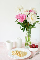 pancakes with strawberries and coffee on the table near a vase with peonies on a white background