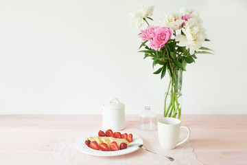 pancakes with strawberries and coffee on the table near a vase with peonies on a white background