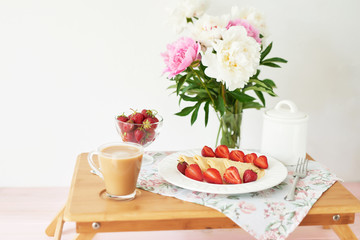 pancakes with strawberries and coffee on the table near a vase with peonies on a white background