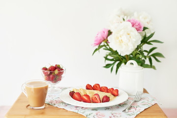 pancakes with strawberries and coffee on the table near a vase with peonies on a white background
