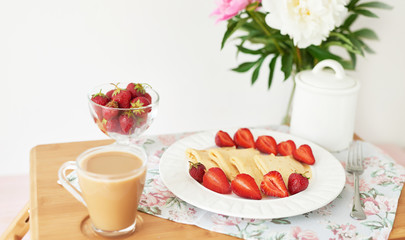 pancakes with strawberries and coffee on the table near a vase with peonies on a white background