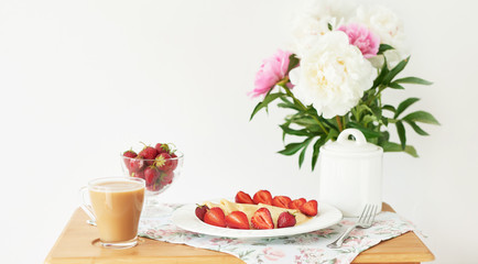 pancakes with strawberries and coffee on the table near a vase with peonies on a white background