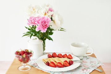pancakes with strawberries and coffee on the table near a vase with peonies on a white background