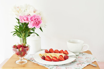 pancakes with strawberries and coffee on the table near a vase with peonies on a white background