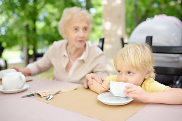 Beautiful senior lady with his little grandson drinking tea in outdoors cafe or restaurant. Elderly lady lifestyle.