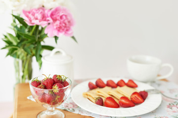 pancakes with strawberries and coffee on the table near a vase with peonies on a white background