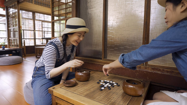 Two beautiful girl friends playing chess with japanese style house interior. young female travelers having fun together with igo go stones. attractive laughing women play chinese board game cheerful