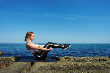 Fototapeta na wymiar Horizontal photo of a beautiful woman who does sports exercises on the beach of the ocean or sea. The girl does exercises of abdominal muscles and holds dumbbells in her hands.