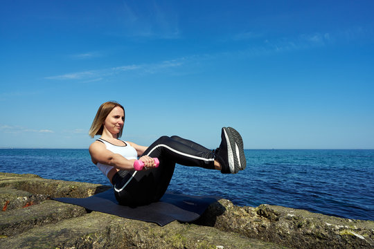 Horizontal photo of a beautiful woman who does sports exercises on the beach of the ocean or sea. The girl does exercises of abdominal muscles and holds dumbbells in her hands.