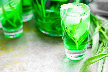 Summer cold green drink with tarragon, mint and lemon in small shot with ice and defocused decanter on background. Sparkling beverage, alcohol drink on dark background. Horizontal, copy space