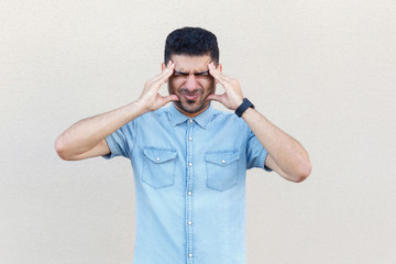 headache, confusion or problem. Portrait of sick handsome young bearded man in blue shirt standing and holding his head and feeling bad. indoor studio shot isolated on light beige wall background.