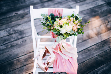 Rustic wedding bouquet and delicate bridal shoes on a white chair.