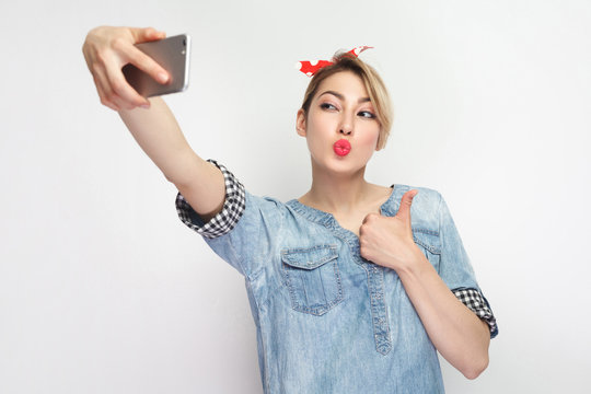 Selfie time! Portrait of sexy attractive blogger woman in casual blue denim shirt with makeup, red headband standing, holding phone and making selfie, showing thumb up and send air kiss. studio shot