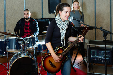 Obraz na płótnie Canvas Attractive female soloist playing guitar and singing with her music band in sound studio