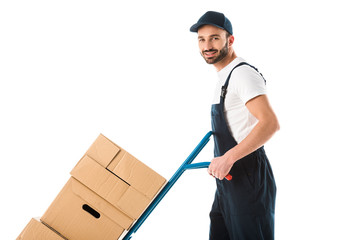 cheerful delivery man transporting hand truck with cardboard boxes and looking at camera isolated...