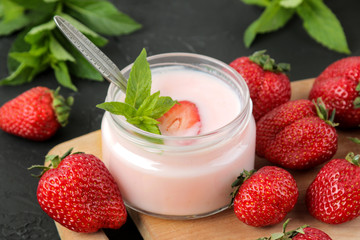 delicious strawberry yogurt in a jar and fresh ripe strawberries and a mint leaf on a black concrete table.