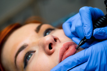 Naklejka premium Permanent make-up for pink lips of beautiful woman in beauty salon. Beautician doing tattooing lips. Micropigmentation. Close Up