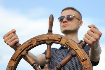 A man controls the yacht, hands on the steering wheel, handwheel on sky background