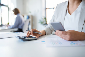 business and people concept - businesswoman with calculator, smartphone and papers working at office