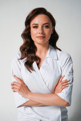 Attractive young female doctor in white medical jacket isolated on white background. Portrait of brunette woman cosmetologist with cross hands
