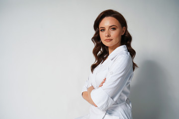 Attractive young professional doctor in white medical jacket isolated on white background. Brunette woman cosmetologist. Cross hands. Space for text. Portrait shot