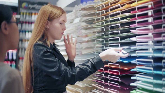 Medium shot of female shop assistant showing colored paper sheets and explaining texture differences to young Asian customer in art store