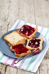 Fresh toasted cereal bread slices with homemade cherry jam and spoon on ceramic plate closeup on linen napkin on rustic wooden table background.