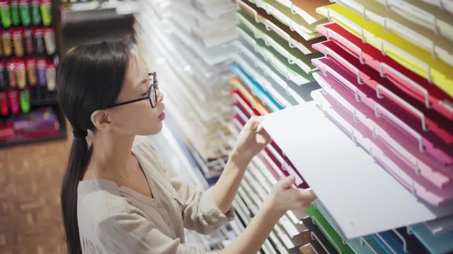 High angle of young Asian artist in eyeglasses selecting white paper among multi-colored sheets placed on holders in art and craft shop