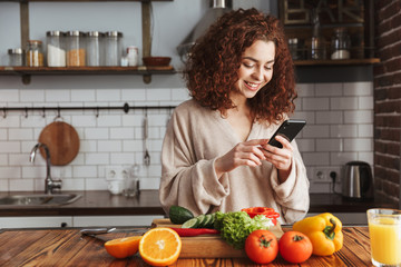 Photo of joyful caucasian woman holding smartphone while cooking salad with fresh vegetables at home
