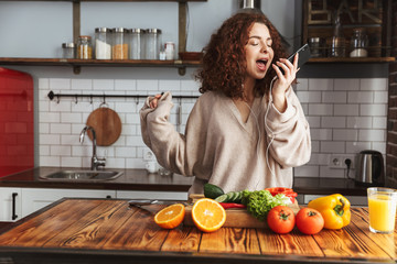 Image of cheery caucasian woman listening to music on mobile phone while cooking in kitchen interior at home