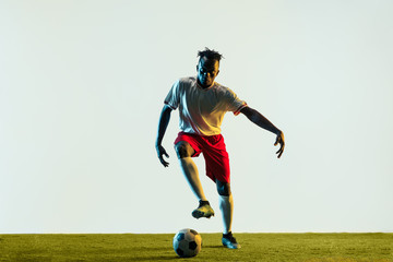 Young african-american male football or soccer player in sportwear and boots kicking ball for the goal in neon light isolated on white background. Concept of healthy lifestyle, professional sport