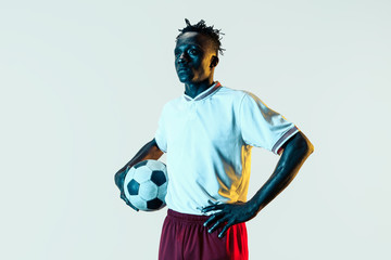 Young african-american male football or soccer player in sportwear and boots standing with the ball in neon light isolated on white background. Concept of healthy lifestyle, professional sport, hobby.