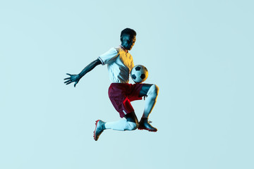 Obraz na płótnie Canvas Young african-american male football or soccer player in sportwear and boots kicking ball for the goal in jump in neon light on gradient background. Concept of healthy lifestyle, professional sport.