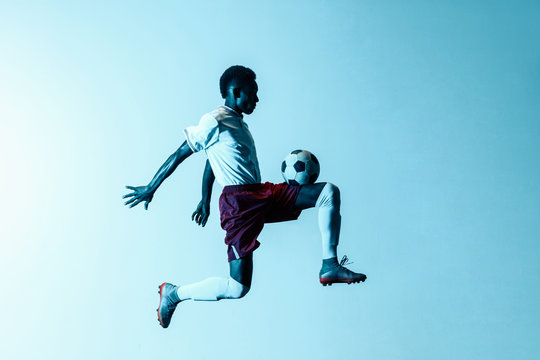 Young african-american male football or soccer player in sportwear and boots kicking ball for the goal in jump in neon light on gradient background. Concept of healthy lifestyle, professional sport.