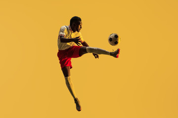 Plakat Young african-american male football or soccer player in sportwear and boots kicking ball for the goal in jump in neon light on gradient background. Concept of healthy lifestyle, professional sport.