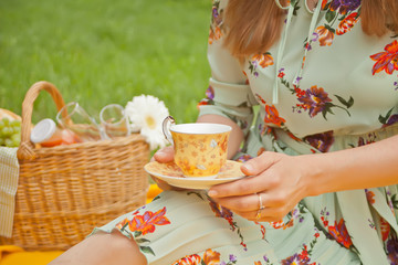 Woman on the picnic sits on the yellow cover and holds cup of tea or coffee. Near picnic basket with food and fruits.