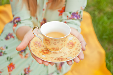 Woman on the picnic sits on the yellow cover and holds cup of tea.