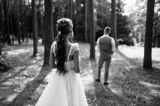 back view of beautiful young bride in wedding dress looking at groom standing in forest, black and white image