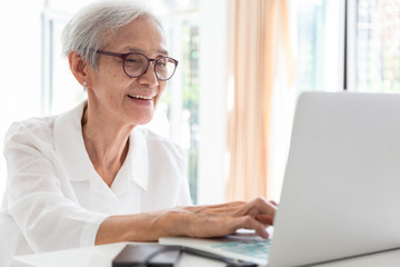 Happy asian senior woman working with laptop computer at table in home,smiling elderly people in glasses with white hair sits in front of the computer and serf web-sites, technology and people concept - Powered by Adobe