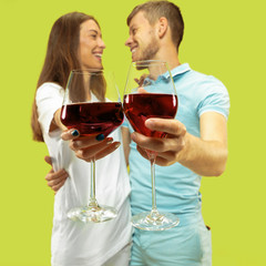 Beautiful young couple's half-length portrait isolated on green studio background. Woman and man standing with glasses of red wine. Facial expression, summer, weekend concept. Trendy colors.