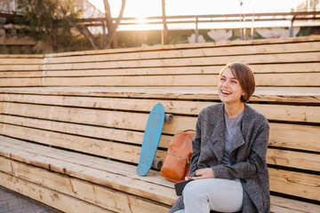 Young laughing woman with a skateboard is sitting on a 2-stage bench.