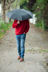 Portrait of man wearing  a red pulllver and blue jeans walking in the street with a black umbrella