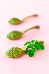 Moringa powder in wooden spoons with original fresh Moringa leaves on pink background.