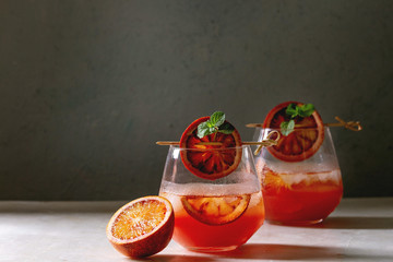 Blood orange iced cocktails in glasses, decorated by slice of oranges and fresh mint on skewers, served on white marble table with grey wall at background.