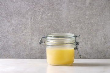 Homemade Melted ghee clarified butter in close glass jar standing on white marble table with grey wall at background. Copy space