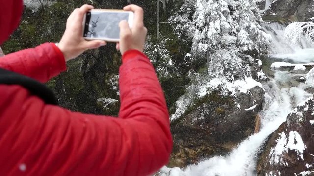 A girl takes pictures of a mountain river in winter. Beautiful winter landscape in the mountains. Red jacket and tourist backpack. Snowy trees in the forest. Active holiday in the mountainous nature