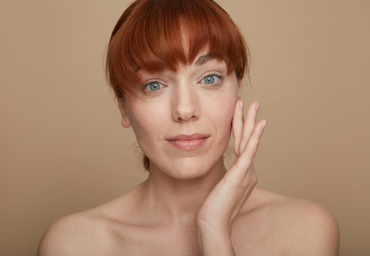 Beautiful red hairered and blue eyes adult woman massaging her face looking at camera over brown background.