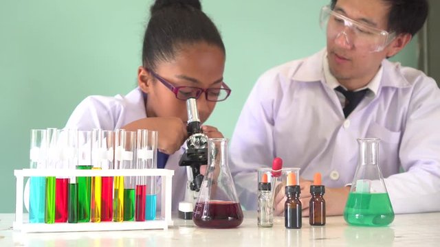 Young Asian scientist teaching African American kid how to use microscope with chemical substance tubes and flasks in classroom - science and learning education concept