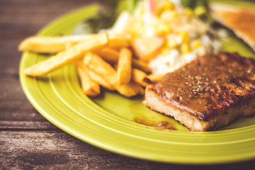 Pig steak is a fast food,pig steak this dish consists of with salad which has  purple lettuce corn apple cabbage salad dressing bread french fries and black pepper sauce, all in a green plate.